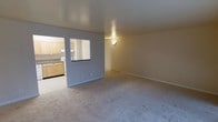 This single-floor unit is designed with a straightforward, functional layout that connects the main living area to the kitchen through both a doorway and a pass-through window. The living area is finished with neutral-toned wall-to-wall carpeting and painted in a light gray-beige palette, creating a clean, understated backdrop. White baseboards run along the perimeter, adding a crisp contrast to the soft flooring and walls. A wall-mounted sconce provides ambient lighting to the room.

The kitchen is accessible through a wide doorway and is visually open to the living space via the pass-through window, enhancing the sense of connectivity between rooms. The kitchen features light wood cabinetry, dark countertops, and tile flooring, offering a practical and functional workspace. White appliances, including what appears to be a range, are integrated into the cabinetry layout. The upper cabinets extend toward the ceiling, maximizing storage capacity.

Overall, the space maintains a neutral, move-in-ready aesthetic with consistent finishes throughout, making it a practical and adaptable living environment.