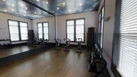 This single-floor fitness studio is thoughtfully designed to support a focused workout environment. The room is equipped with several stationary spin bikes arranged on a warm hardwood floor, providing ample space for cycling sessions. A full-length mirror spans one wall, amplifying the sense of space and allowing users to monitor their form during exercise. Along the opposite wall, a rack of kettlebells and additional strength training equipment offers supplementary workout options beyond cycling. Natural light filters into the room through multiple windows fitted with horizontal blinds, contributing to a bright and energizing atmosphere. One of the most distinctive design elements is the ceiling, which features a dramatic painted sky mural complete with clouds, accented by recessed spotlights that add a dynamic, moody ambiance to the space. The dark window trim and neutral wall tones complement the overall aesthetic, creating a sleek, modern feel. This well-appointed studio balances functional fitness equipment with thoughtful design details, making it suitable for both individual training and small group fitness classes.