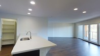 This single-floor space is designed with an open-concept layout that promotes a sense of airiness and connectivity between the kitchen and living areas. The kitchen is anchored by a clean, white countertop island equipped with a stainless steel sink, offering both functional workspace and a visual centerpiece. Dark-toned hardwood flooring runs throughout the space, providing a striking contrast against the bright white walls and cabinetry, adding warmth and a contemporary aesthetic. Recessed lighting is thoughtfully installed across the ceiling, ensuring consistent, well-distributed illumination throughout the entire floor plan. The living area is generously proportioned, featuring neutral white walls that offer a versatile canvas for a variety of interior design styles. Electrical outlets are conveniently placed along the walls, catering to practical needs. The minimalist design and clean lines throughout the space reflect a modern sensibility, making it an ideal setting for those who appreciate an uncluttered, functional environment. The seamless transition between the kitchen and living areas enhances the overall flow, making the space feel expansive and welcoming.