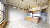 This two-floor space showcases a striking industrial aesthetic defined by its exposed concrete ceiling, where visible ductwork and conduit run overhead, adding an authentic urban character. The polished concrete floors throughout the main level complement the raw ceiling, creating a cohesive industrial look balanced by contemporary touches.

The open-concept layout centers around a well-appointed kitchen featuring warm wood cabinetry in a rich medium-brown tone, paired with a large kitchen island finished in matching wood with a light-colored countertop surface. The island serves as both a functional workspace and a natural divider between the kitchen and the open living area. Stainless steel appliances, including a full-size refrigerator, are integrated into the cabinetry, contributing to a modern, streamlined appearance.

Pendant lighting with geometric cage-style fixtures hangs above the island, bridging the industrial and contemporary design elements. The spacious open living area offers a generous, flexible footprint, while neutral gray walls provide a clean contrast to the textured ceiling above. The two-floor configuration adds further dimension and versatility to this distinctive urban space.