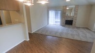 This single-story space offers an open and functional layout that seamlessly combines a dining or entry area with a spacious living room. The transition between the two zones is clearly defined by a shift from rich hardwood-style flooring near the entry to soft, neutral-toned carpet throughout the living area, adding warmth and visual interest to the overall design.

The living room is anchored by a prominent brick fireplace, which serves as a natural focal point and adds a classic, cozy character to the space. A large mirror is mounted above the fireplace, enhancing the sense of openness and reflecting light throughout the room. Vertical blinds frame a full sliding glass door, which allows for generous natural light and provides access to an outdoor area.

Overhead, a ceiling fan with a light fixture provides both comfort and illumination, complementing the neutral beige wall tones that keep the space feeling clean and inviting. A partial wall or bar divider near the entry adds subtle architectural definition without disrupting the open flow between areas. Overall, the space presents a comfortable, well-lit environment with practical features and timeless design elements.