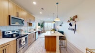 This single-floor residence showcases a thoughtfully designed open-concept layout that seamlessly connects the kitchen, dining, and living areas into one cohesive space. The kitchen is outfitted with warm wood cabinetry, white subway tile backsplash, stainless steel appliances including a side-by-side refrigerator, electric range, built-in microwave, and dishwasher. A large central island with a white quartz countertop doubles as a dining surface, complemented by stylish bar seating. Pendant lighting overhead adds a modern mid-century touch. Light wood-style flooring runs throughout, enhancing the bright, airy atmosphere created by recessed lighting and large windows that draw in natural light. A bold teal entry door adds a striking accent upon arrival.

The living area extends naturally from the kitchen, offering generous space and outdoor views through expansive windows. Both bedrooms are comfortably sized with light-filled windows and the same warm flooring. The bathroom features white subway tile surrounding a full tub-shower combination, a wide single-sink vanity with white cabinetry and quartz countertop, and a large mirror. Clean lines and a neutral palette unify the space with a contemporary, welcoming ambiance.