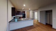 This single-floor unit offers a well-organized layout that balances functional living spaces with a clean, modern aesthetic. The kitchen serves as a central highlight, outfitted with rich, dark espresso cabinetry that extends to the ceiling, providing ample storage throughout. Black appliances, including a range, built-in microwave, and refrigerator, complement the dark granite-style countertops. Track lighting illuminates the workspace, while a peninsula with a double-basin sink and extended counter space adds convenience and delineates the kitchen from the adjoining living area. The flooring transitions from warm hardwood-style planks in the kitchen and living spaces to plush carpet in the bedroom. The bedroom is a comfortable, quiet space featuring neutral walls and a large double-hung window that admits natural light and offers views of the surrounding streetscape. The full bathroom is finished with large-format dark gray tile flooring, a white tub-shower combo with integrated shelving, a toilet, and a dark vanity, maintaining a cohesive, contemporary palette. The overall layout, visible in the overhead floor plan, flows efficiently between rooms, with clean white walls and recessed lighting throughout contributing to a bright, functional atmosphere.