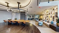 This thoughtfully designed single-floor amenity space combines a polished lobby with a multifunctional lounge and coworking environment. The grand entrance features soaring double-height ceilings, large floor-to-ceiling glazed walls that flood the space with natural light, light-toned large-format tile flooring, and a striking open staircase with glass railings and dark metal accents. Potted greenery and curated black-and-white artwork add warmth to the otherwise crisp entryway.

Transitioning deeper into the space, rich dark hardwood flooring contrasts elegantly with the lighter lobby tones. A sculptural, oversized circular ring pendant light fixture commands attention overhead, suspended above a long communal dining or conference table outfitted with tan leather chairs. The ceiling features curved recessed lighting details that define zones within the open layout.

A generous lounge area anchors one side of the space, furnished with plush sofas, light blue accent chairs, and a neutral area rug, all set against a floor-to-ceiling illuminated shelving unit filled with books and décor. Adjacent to the lounge, a glass-walled coworking room provides a semi-private workspace. A digital display panel and elevator access are also integrated seamlessly into the design.