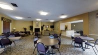 This well-appointed party room offers a welcoming and functional space designed to accommodate a variety of social gatherings and events. The main area is furnished with multiple round tables and cushioned chairs, providing ample seating for guests throughout the open floor plan. Warm yellow-toned walls contribute to an inviting atmosphere, complemented by patterned carpeting that adds texture and comfort underfoot. Recessed flush-mount ceiling light panels ensure the space is evenly and brightly illuminated throughout.

A cozy lounge area anchors one end of the room, centered around a classic brick fireplace and framed by comfortable seating — perfect for more relaxed conversation. Decorative potted plants and framed artwork are placed throughout, adding warmth and a finished aesthetic to the environment.

Adjacent to the main gathering area, a connected kitchenette or service counter is visible through an open pass-through, outfitted with pendant lighting and counter space, making it convenient for catering or refreshment setup. A wall-mounted television is also present, adding an entertainment option for events. Overall, the space balances practicality with comfort, making it a well-rounded venue for community or private functions.