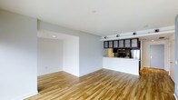 Located in Dallas, this single-floor space offers a clean, modern aesthetic with an open-concept layout that maximizes the sense of space and functionality. Warm-toned hardwood flooring runs throughout, providing a welcoming contrast to the crisp white walls and ceilings that lend the interior a bright, airy feel.

The living area is spacious and open, seamlessly connected to the kitchen through a raised bar-height counter that doubles as a casual dining or entertaining surface. The kitchen is outfitted with sleek dark-framed cabinetry featuring glass-panel upper doors, stainless steel appliances including a refrigerator and microwave, and a tile backsplash that adds a touch of visual texture to the otherwise minimalist palette.

Recessed lighting is integrated into the ceiling, supplemented by a track lighting system positioned near the kitchen, ensuring the space is well-lit throughout. The overall design embraces a contemporary urban sensibility — clean lines, neutral tones, and thoughtful detailing come together to create a versatile environment suitable for both relaxed daily living and stylish entertaining. The open floor plan and cohesive design make this Dallas space both functional and visually appealing.