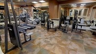This fitness center offers a comprehensive workout environment designed to accommodate a wide range of exercise needs. The space is outfitted with an extensive selection of cardio equipment, including treadmills and an elliptical trainer, providing ample options for cardiovascular training. A variety of strength and resistance machines are thoughtfully arranged throughout the room, catering to full-body workouts for users of all fitness levels. Floor-to-ceiling mirrors line the walls, creating a sense of openness while allowing users to monitor their form during exercise. The warm, earth-toned walls contribute to an inviting and energizing atmosphere, complemented by the distinctive patterned carpet that adds a touch of elegance to the functional space. Overhead lighting ensures the entire area is well-illuminated, and a wall-mounted clock and television screen add practical and entertainment value. A dedicated free weights and bench area rounds out the facility, making this fitness center a fully equipped destination for a complete and varied workout experience.