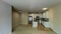 This single-floor space features an open-concept layout that seamlessly connects the kitchen and living area. The kitchen is well-appointed with white cabinetry extending to the upper walls, dark granite countertops, and a full suite of stainless steel appliances, including a refrigerator, range, and built-in microwave. A decorative tile backsplash with a mosaic pattern adds visual interest along the cooking wall. A central island with matching dark countertop provides additional workspace and serves as a natural dividing point between the kitchen and the adjoining living space. The sink is positioned along the far wall beneath additional cabinetry, offering ample storage throughout. Track lighting runs across the ceiling in both the kitchen and living areas, providing adjustable, directional illumination. The living area features wall-to-wall carpet in a neutral tone, contrasting with the hardwood-style flooring in the kitchen. The walls are painted in a soft, neutral beige, contributing to a clean and cohesive aesthetic. Overall, the space offers a functional, modern design suited for everyday living.