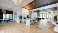 Located at 4535 SW 54th St Rd in Ocala, Florida, the leasing office at Darley Luxury Apartments is a thoughtfully designed, open-concept space that immediately conveys a sense of upscale living. Light hardwood flooring with teal accent inlays runs throughout, complementing a sophisticated palette of white, dark charcoal, and warm wood tones. The space is anchored by a prominent white reception and bar-style counter complete with gray upholstered seating, ideal for welcoming prospective residents. Above, a striking chandelier and a grid of pendant drop lights draw the eye upward, while warm woven rattan pendant fixtures illuminate the lounge areas beyond. A multi-zone layout separates the reception area from two distinct lounge spaces. One features a bold teal sofa set against a dark accent wall adorned with a chevron-patterned living moss installation, flanked by abstract line-art wallpaper and floor-to-ceiling windows that invite natural light. The other seating area offers plush white chairs, curated artwork, and textured stone-clad accent walls. The Darley Luxury Apartments branding is prominently displayed in gold lettering, reinforcing the premium character of the community. A dedicated glass-fronted leasing office provides a private area for consultations, completing this polished and welcoming environment.