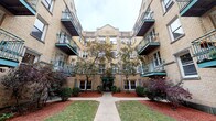 This residence is situated within a distinguished multi-story courtyard building, characterized by its warm yellow brick facade, ornamental detailing, and teal-railed balconies that frame a landscaped central courtyard. A welcoming green canopy marks the building entrance, flanked by mature trees and well-maintained shrubbery, reflecting the architectural charm typical of early 20th-century Chicago construction.

Inside, the single-floor unit features warm hardwood flooring throughout the main living areas and bedrooms, contributing to a cohesive and inviting atmosphere. The open living area is equipped with a ceiling fan and connects directly to the kitchen, which offers ample light-wood cabinetry, a tiled backsplash, corner sink, gas range, and full-size refrigerator — providing a practical and well-organized cooking space.

Two bedrooms are present, each with hardwood floors, ceiling fans or windows that bring in natural light, and classic cast-iron radiators for heating. The bathroom features white tile throughout, a soaking tub with shower, a vanity with sink, and a frosted window for natural ventilation. The unit's clean, neutral palette and traditional details create a functional and comfortable living environment.