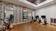 Located at 2180 Pickens Street in Columbia, SC, this well-appointed fitness studio offers a thoughtfully designed space for cardiovascular and strength training. The room is defined by a striking botanical and bird-patterned accent wall that adds a bold, artistic character to the environment, setting it apart from a typical gym aesthetic. A tray ceiling with recessed and pendant lighting creates a sense of height and architectural interest, while the warm cork-style tile flooring provides a durable yet visually inviting surface underfoot. Multiple Peloton-style spin bikes, each equipped with individual display screens, line the room and serve as the centerpiece of the cardio offerings. A large wall-mounted flat-screen television provides an additional focal point for group or guided workouts. Full-length arched mirrors with sleek black frames are strategically placed to enhance the sense of space and support proper form during exercise. A well-stocked dumbbell rack and an assortment of medicine balls and stability balls round out the strength and functional training options. Tall glass double doors with dark frames open to an adjacent area, allowing natural light to filter in and connecting the studio to the broader facility. The overall ambiance is energetic yet refined, blending functional fitness amenities with intentional interior design.