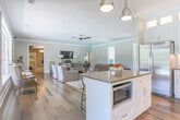 Located in Columbus, Georgia, this single-floor space combines classic Southern charm with thoughtfully modern interior design. Rich hardwood flooring flows seamlessly throughout, anchoring each area with warmth and continuity. Crown molding and crisp white trim add an elegant architectural finish to the neutral gray walls, creating a polished yet inviting atmosphere.

The expansive living area is generously proportioned, offering multiple seating arrangements centered around a rustic wooden coffee table atop a patterned area rug. Soft, upholstered sofas and accent chairs invite comfortable gathering, while large multi-pane windows frame lush outdoor greenery and pool views, filling the room with natural light. A wall-mounted flat-screen TV and a distressed wood media console add functional style to the space.

The kitchen is a standout feature, showcasing floor-to-ceiling white Shaker-style cabinetry with glass-panel upper doors and under-cabinet lighting. A stone tile backsplash, quartz countertops, and stainless steel appliances complete the refined aesthetic. A central island with bar seating and industrial-style pendant lighting makes the kitchen both practical and visually striking. A glass-paned exterior door provides direct outdoor access and additional natural light. The overall ambiance is calm, cohesive, and elegantly appointed.