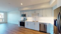 Located in Los Angeles, CA, this modern single-story studio presents a thoughtfully designed open-concept layout that seamlessly integrates living and kitchen areas. The space is finished with warm-toned hardwood flooring that runs throughout, complementing the bright, neutral color palette and recessed lighting overhead.

The kitchen is a standout feature, outfitted with vertical shaker-style cabinetry in a soft light gray finish, extending floor-to-ceiling for generous storage. Clean white countertops provide ample prep space, while a full suite of stainless steel appliances — including an electric range, over-the-range microwave, dishwasher, and French door refrigerator — adds a polished, modern touch. A single-basin sink with a contemporary faucet completes the functional layout.

The open living area benefits from natural light streaming through large windows that frame views of the surrounding Los Angeles neighborhood, creating an airy and inviting atmosphere. A separate room offers versatility as a bedroom or workspace, with its own window providing additional natural light and outdoor views.

The bathroom features a soaking tub with tiled surround, a white undermount sink set into a gray vanity, a large mirror, and light tile flooring, maintaining the clean, contemporary aesthetic found throughout the rest of the space.