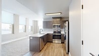This single-floor residence offers a thoughtfully designed layout that balances modern finishes with a welcoming, open feel. The kitchen is a standout feature, showcasing rich dark-toned cabinetry, a full suite of stainless steel appliances including a range, microwave, dishwasher, and refrigerator, and a peninsula with a white countertop and undermount sink. Warm wood-look flooring runs throughout the kitchen and connecting areas, lending a cohesive, contemporary aesthetic.

The living and sleeping areas are finished with plush neutral-toned carpet and painted in soft greige tones that create a calm, understated ambiance. Multiple large windows line the walls, flooding the rooms with natural light and offering impressive elevated views of the surrounding cityscape and skyline — a defining characteristic of this upper-floor residence. The generous ceiling height enhances the sense of openness throughout.

Bedrooms provide ample space, with one featuring a sliding closet and additional windows with roller shades for light control. A bathroom with wood-look flooring, a white vanity, and matte black hardware accents completes the unit. The overall design is clean and modern, combining functional layouts with quality finishes suited for urban living.