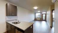 This single-floor unit features an open-concept layout that integrates the kitchen and living area into one cohesive, functional space. The kitchen is equipped with granite countertops speckled in neutral tones, a double-basin stainless steel sink, dark espresso-finished cabinetry, and a built-in dishwasher. The peninsula-style counter serves as a natural divider between the kitchen and the living area, making the space feel connected while maintaining distinct functional zones.

The living area extends generously from the kitchen, finished with durable hardwood-style flooring in a warm, medium-brown tone that runs throughout the unit. White walls and flush-mount ceiling light fixtures keep the space feeling clean and contemporary. Natural light enters through a large window dressed with grey curtains, and a glass-panel door provides access to what appears to be a private balcony, further enhancing the sense of openness. The overall ambiance is modern and well-maintained, with a neutral palette that offers flexibility for a variety of interior styles.