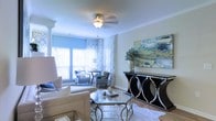 This single-floor space showcases a refined interior design with a carefully curated aesthetic that blends comfort and elegance. The living area serves as the centerpiece of the home, bathed in natural light that streams through large windows dressed with patterned blue and white curtains, complemented by classic wooden blinds. Crown molding frames the ceiling, which features a ceiling fan with a frosted glass light fixture, adding a polished finishing touch overhead. The space is anchored by a plush neutral sofa and a sleek glass-top coffee table with a gilded metal frame, creating a sophisticated yet approachable seating arrangement. A set of upholstered accent chairs in muted blue tones are positioned near the sun-lit corner, offering an inviting reading or conversation nook. A striking black and chrome console table along the wall displays decorative accessories including a lush succulent arrangement and an eye-catching abstract painting in shades of blue and gold, tying the room's color story together. Light hardwood flooring runs throughout, enhancing the open, airy ambiance of the space. The overall design balances contemporary elegance with warm, livable comfort.