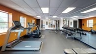 This single-floor space is designed with a cohesive modern aesthetic, anchored by a striking color palette of orange and neutral tones throughout. The lobby entrance makes an immediate impression with its large-format white tile flooring, a distinctive orange ceiling feature with a decorative chandelier, and floor-to-ceiling glass walls that flood the space with natural light and offer views of surrounding greenery. An elevator and secured access points are conveniently located within the lobby, reinforcing the functional layout.

Adjacent to the entrance, a lounge area is furnished with tasteful high-back chairs and side tables, positioned alongside expansive glass walls that frame outdoor views of mature trees and landscaping. Curated artwork and layered rugs add a refined, welcoming character to the seating area.

The fitness center is a well-appointed, generously sized room featuring rubber flooring, a drop tile ceiling with panel lighting, and bold orange accent walls. It is equipped with cardio machines including treadmills, ellipticals, and a stationary bike, as well as a full dumbbell rack and weight equipment. Floor-to-ceiling mirrors expand the sense of space. A small transitional lounge area with decorative seating connects the fitness center to the broader space, providing a thoughtful buffer between activity zones.