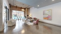 Located at 201 Jefferson St N in the heart of downtown Huntsville, Alabama, this single-floor amenity space masterfully combines industrial character with contemporary design. Polished concrete floors extend throughout, reflecting light and lending a sleek, cohesive feel to the entire space.

The main lounge area is anchored by exposed brick columns and walls that contrast beautifully against crisp white painted surfaces and a painted concrete ceiling with visible ductwork — hallmarks of an industrial-modern aesthetic. Generous floor-to-ceiling glass windows frame views of an outdoor pool area, flooding the interior with abundant natural light. The lounge is furnished with a large sectional sofa, accent chairs, and a central coffee table arranged atop a patterned area rug, creating a welcoming social environment. Colorful artwork adorns the walls, adding warmth and visual interest.

Adjacent to the lounge, a bar and kitchenette area features rich wood cabinetry, granite countertops, bar seating, pendant lighting, and mounted flat-screen televisions set against a textured tile backsplash — ideal for gatherings and events.

A fully equipped fitness center is prominently visible through full-height glass walls, housing a variety of strength and cardio equipment. The transparent partitions maintain an open, connected atmosphere while defining the dedicated workout space. Together, these areas offer a well-appointed and functional communal environment.