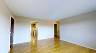 This single-floor residence offers a straightforward and functional layout designed for comfortable everyday living. The main living area is characterized by smooth hardwood flooring in a warm honey tone, which adds a welcoming brightness to the space. Clean white walls and a simple white ceiling contribute to an open, airy ambiance that makes the area feel well-lit and inviting. From the living area, an open doorway leads directly into the kitchen, which features wood-toned cabinetry and standard appliances, providing a practical and accessible cooking space. A second doorway off the living area opens to an additional room, finished with a deep green accent wall that provides a distinct visual contrast to the neutral tones of the main space. Baseboards run cleanly along the walls throughout, and the overall design maintains a consistent, understated aesthetic. The single-floor plan ensures easy navigation between the living, cooking, and adjoining spaces, making the layout efficient and cohesive. The space presents a clean canvas suitable for a variety of interior styling preferences.