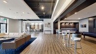 This contemporary single-floor amenity space offers a generously sized, open-plan layout designed for socializing and recreation. The space is divided into several well-defined functional zones that flow seamlessly into one another, all unified by warm wood-tone luxury vinyl flooring throughout.

The lounge area anchors one end of the space, featuring a sleek linear gas fireplace with a mounted flat-screen TV above, cream-toned sectional sofas, and a bold navy herringbone-patterned area rug. Floor-to-ceiling windows flood this zone with natural light and offer views of the surrounding streetscape.

Adjacent to the lounge, a game and entertainment zone includes a pool table, dartboard, high-top bar tables with modern barstools, and built-in dark wood banquette seating beneath recessed accent lighting — creating a lively, lounge-bar atmosphere.

The kitchen and dining area features dark cabinetry, a stainless steel refrigerator, open shelving, and multiple high-top communal tables with contemporary seating, ideal for casual dining or gatherings. Large windows provide ample natural light in this zone as well.

Overhead, an exposed black ceiling with visible ductwork and industrial-style track lighting adds an urban, loft-inspired contrast to the polished, modern finishes throughout the rest of the space. The overall ambiance is refined yet inviting, balancing comfort with a distinctly contemporary aesthetic.