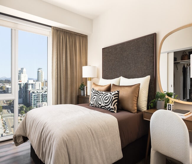 Chic and cozy bedroom featuring a plush bed with decorative pillows, elegant desk with mirror, and a stunning city view through large windows. - Ancora Apartments
