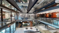 Located at 4545 Laclede Ave in St. Louis, Missouri, this thoughtfully designed commercial space makes an immediate impression with its dramatic double-height atrium, framed by glass railings and dark structural beams. A sweeping large-scale abstract mural anchors the central volume, visible from multiple vantage points and serving as a bold artistic focal point throughout the space.

The upper level opens onto curated lounge areas featuring curved sectional seating, statement pendant lighting, and warm-toned accent walls adorned with ornate mirrors — creating an inviting, upscale atmosphere. Vertical wood slat panels appear throughout, adding warmth and texture that contrasts elegantly with the darker gray walls and polished surfaces found in the corridors and transitional areas.

A dedicated lounge zone with deep navy banquette seating, terracotta-hued walls, and a geometric brass ceiling fixture offers a rich, intimate setting. Private meeting rooms feature natural light filtered through sheer copper-toned drapes, warm wall paneling, and pendant globe lighting, making them well-suited for focused gatherings.

Sleek hallways clad in brushed charcoal finishes connect the various zones, lending a refined industrial edge. Throughout the space, the design balances contemporary sophistication with artistic expression, creating a dynamic and functional environment ideal for professional and hospitality use.