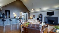 This single-story home showcases a thoughtfully designed open-concept layout that connects the main living areas into one cohesive space. The living room is anchored by dramatic vaulted ceilings that rise to a sharp peak, enhancing the sense of volume and airiness throughout. A ceiling fan is centrally mounted, providing both function and a finishing architectural detail.

The living area is furnished with rich brown leather sofas and Mission-style wood accent tables, lending a warm, grounded aesthetic. Hardwood flooring runs throughout, adding continuity and warmth to the space. A bookcase along one wall provides storage and display space, while framed artwork adds character to the dark-painted accent walls.

Transitioning seamlessly from the living room, the kitchen and breakfast bar area is visible in the background, featuring bar-height seating and a well-appointed counter setup. The open sightlines between rooms make the entire floor plan feel expansive and interconnected.

Natural light filters through the space, complementing the recessed lighting and wall-mounted fixtures. The overall ambiance is casual yet refined, with a cohesive color palette of deep grays, warm browns, and natural wood tones throughout.
