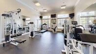 Located at 3539 Mary Taylor Rd in Birmingham, Alabama, this fitness center offers a well-appointed, single-floor workout facility designed to accommodate a full range of exercise needs. The space is characterized by its bright, welcoming atmosphere, with crisp white walls, crown molding, and durable rubber flooring throughout that provides both comfort and functionality underfoot.

The main workout area is generously sized and houses an array of strength training machines, including Paramount cable and resistance units, as well as free weight equipment and adjustable benches. Cardio offerings include treadmills and a stationary recumbent bike, all positioned in front of large windows that allow natural light to flood the space and provide views of the surrounding outdoor environment. Multiple wall-mounted televisions are strategically placed throughout, offering entertainment during workouts.

A dedicated stretching and functional training alcove is also present, featuring a more intimate setting with decorative wall shelving, yoga mats, and a flat bench — ideal for warm-ups, cool-downs, or focused training sessions.

Elegant French doors connect the various areas of the facility, enhancing the open flow between spaces. High ceilings with crown molding trim lend an upscale feel uncommon in fitness centers, while recessed and flush-mount lighting ensures the space remains well-lit throughout. The overall ambiance is clean, organized, and inviting.