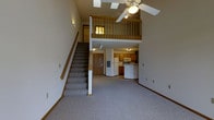 This two-story residence offers a well-designed layout that maximizes both space and functionality. The main level features an open-concept living and kitchen area, where the generous ceiling height creates an airy, expansive atmosphere. A ceiling fan with integrated lighting is positioned overhead, enhancing comfort throughout the living space, which is finished with wall-to-wall carpet and clean neutral-toned walls.

The kitchen is positioned at the rear of the main level and is equipped with wooden cabinetry, a white refrigerator, and a built-in microwave and range. A peninsula-style counter provides additional workspace and helps define the boundary between the kitchen and the living area. A wood-paneled entry door is visible adjacent to the kitchen, offering convenient access.

A carpeted staircase with a warm wooden handrail leads to the upper loft level, which overlooks the main living area through an open wooden railing. This elevated space benefits from the dramatic ceiling height and is illuminated by a skylight, flooding the area with natural light. The two-level design of this residence creates a dynamic and versatile living environment suited for a variety of lifestyle needs.