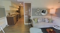 This single-floor space showcases a thoughtfully designed open-concept layout that seamlessly integrates the kitchen and living area. The kitchen is equipped with stainless steel appliances, including a refrigerator and range, complemented by sleek cabinetry in a warm wood tone paired with white upper cabinets. A stainless steel sink with modern fixtures adds a polished touch, while recessed lighting illuminates the space efficiently.

A striking frosted glass partition element serves as a visual divider between the kitchen corridor and the living area, allowing light to pass through while providing subtle definition between spaces. The living area features a light-toned sofa accented with colorful decorative pillows in green and patterned designs, anchored by a dark area rug. A small black side table provides functional accent furniture.

The accent wall in the living area is finished in a muted grey-green hue with a horizontal stripe detail, adorned with a curated arrangement of round decorative mirrors of varying sizes. Light wood-look flooring runs throughout, unifying the space and enhancing its warm, contemporary ambiance. Natural light appears to enter through a window or glass door visible in the background.