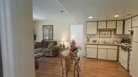 This single-floor residence offers a welcoming and functional open-concept layout that integrates the living, dining, and kitchen areas into one cohesive space. Warm hardwood flooring flows throughout, lending a sense of continuity and warmth to the interior.

The living area is comfortably appointed, featuring a plush sofa accented with decorative pillows, a classic table lamp, and wall art that adds character to the neutral, softly painted walls. A textured ceiling adds subtle architectural interest overhead.

Adjacent to the living space, the dining area centers around a round glass-top table paired with woven bistro-style chairs — a practical yet charming spot for casual meals. The open sightlines between these areas create an airy, connected feel.

The kitchen is efficiently laid out with white cabinetry, light countertops, and a sink positioned near a window that allows natural light to filter in. Upper cabinetry with warm-toned trim adds a traditional touch to the space.

A hallway visible from the main living area leads to additional rooms beyond, suggesting a thoughtfully organized floor plan. Overall, the space presents a bright, tidy, and comfortable environment with a classic residential aesthetic.