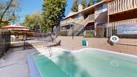 Located at 10824 Fair Oaks Blvd in Fair Oaks, California, this outdoor amenity space centers around a generously sized swimming pool and an adjacent spa, set within a secure, fenced enclosure. The pool features clear, blue-green water and is accessible via built-in stainless steel entry rails, with depth markers along the perimeter indicating a gradual range suitable for a variety of swimmers. A dedicated spa area sits adjacent to the main pool, offering a more intimate setting for relaxation.

The pool deck is finished with a clean concrete surround and includes lounge seating, providing a comfortable space for sunbathing and leisure. A covered carport-style structure along one side offers shaded seating and partial shelter from the sun. The entire area is enclosed by wrought iron fencing, ensuring both safety and privacy.

Towering mature trees frame the space on multiple sides, lending a natural, serene atmosphere and offering additional shade throughout the day. The surrounding multi-story residential buildings, clad in warm wood-toned siding with balconies and staircases, contribute to the cohesive character of the community. Under clear California skies, this outdoor space presents an inviting, well-appointed retreat for residents seeking relaxation and recreation.