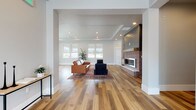 This single-floor space is designed with a modern aesthetic, showcasing an open-concept layout that seamlessly connects the living area and kitchen. Wide-plank hardwood flooring in warm natural tones runs throughout, providing a cohesive and inviting foundation.

The living area is a standout feature, anchored by a floor-to-ceiling fireplace that combines exposed brick with gray shiplap paneling, creating a striking focal point. A tray ceiling with recessed lighting adds architectural depth overhead. Built-in shelving with gray cabinetry flanks the fireplace, offering both display and storage functionality. Multiple windows fitted with roller shades allow ample natural light to fill the space while maintaining privacy.

The kitchen is equally well-appointed, featuring gray shaker-style cabinetry that extends to the ceiling, with glass-front upper cabinets adding visual interest. White quartz countertops contrast cleanly against the cabinetry, and a white subway tile backsplash reinforces the modern palette. A central island provides additional workspace and storage. Stainless steel appliances, including a refrigerator and built-in microwave, complete the kitchen's functional design.

An entry area with a console table provides a transitional space, and the open sightlines throughout ensure the overall ambiance remains bright, spacious, and cohesive.
