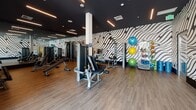Located at 205–233 E Seaside Way in Long Beach, California, this contemporary fitness center delivers a well-appointed workout environment designed for a comprehensive exercise experience. The single-floor facility is defined by its striking black-and-white wave-pattern wall murals, which span multiple walls and lend a bold, energetic character to the space. Dark, recessed ceilings with linear LED lighting fixtures create a sleek, modern atmosphere throughout.

The strength training area is generously equipped with a full rack of dumbbells, multiple cable and selectorized weight machines, adjustable benches, and resistance accessories including bands, stability balls, and yoga mats stored on dedicated racks. Full-length mirrors line one wall, enhancing the sense of space and allowing users to monitor their form during workouts. Wood-tone flooring runs throughout, adding warmth against the darker ceiling palette.

A dedicated cardio zone features a row of Matrix treadmills and elliptical trainers positioned along floor-to-ceiling glass windows, offering natural light and open outdoor views — an especially inviting feature given the facility's proximity to the Long Beach waterfront. The overall layout is open and spacious, with clearly defined zones for both cardio and strength training, making this fitness center a well-rounded and visually dynamic space for exercise.