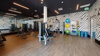 Located at 205–233 E Seaside Way in Long Beach, California, this contemporary fitness center delivers a well-appointed workout environment designed for a comprehensive exercise experience. The single-floor facility is defined by its striking black-and-white wave-pattern wall murals, which span multiple walls and lend a bold, energetic character to the space. Dark, recessed ceilings with linear LED lighting fixtures create a sleek, modern atmosphere throughout.

The strength training area is generously equipped with a full rack of dumbbells, multiple cable and selectorized weight machines, adjustable benches, and resistance accessories including bands, stability balls, and yoga mats stored on dedicated racks. Full-length mirrors line one wall, enhancing the sense of space and allowing users to monitor their form during workouts. Wood-tone flooring runs throughout, adding warmth against the darker ceiling palette.

A dedicated cardio zone features a row of Matrix treadmills and elliptical trainers positioned along floor-to-ceiling glass windows, offering natural light and open outdoor views — an especially inviting feature given the facility's proximity to the Long Beach waterfront. The overall layout is open and spacious, with clearly defined zones for both cardio and strength training, making this fitness center a well-rounded and visually dynamic space for exercise.