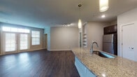 This single-floor residence is designed with a cohesive, modern aesthetic throughout. The open-concept kitchen and living area serves as the heart of the space, anchored by a granite-topped island with an undermount sink and a sleek arc faucet. Dark espresso cabinetry lines the kitchen walls, complemented by stainless steel appliances including an electric range, over-the-range microwave, and refrigerator. Pendant lighting illuminates the island and entryway, while recessed and track lighting provide additional coverage. Rich, dark hardwood-style flooring runs throughout the main living and kitchen areas, creating a warm and polished foundation. Built-in open shelving in the living area adds a functional architectural detail.

The bedrooms are finished with plush carpet and feature ceiling fans, natural light from window blinds, and generous closet space — including at least one walk-in closet with multi-tier shelving. Two bathrooms continue the granite and dark cabinetry theme; one includes a soaking tub with large-format tile surround, while the other features a glass-enclosed walk-in shower with tile walls. Both bathrooms include wood-style flooring, framed mirrors, and wall sconce lighting, maintaining a consistent, refined finish.