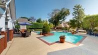 Located at 7827 N Jefferson Place Cir in Baton Rouge, Louisiana, this single-story community facility offers an inviting retreat defined by its classic Southern architectural style. The building features a warm tan and brick exterior, complemented by a gabled roofline, arched windows, and decorative shutters that lend a charming, residential character to the space.

The centerpiece of this property is its expansive outdoor swimming pool, which commands attention with its clear turquoise water and generous dimensions. The pool area is thoughtfully designed, featuring brick-trimmed raised planters with manicured topiary accents that add a sculptural, upscale touch. A broad concrete pool deck surrounds the water, furnished with multiple lounge chairs and a shaded seating area complete with a large patio umbrella — ideal for relaxing outdoors.

The grounds are enclosed by a black iron fence and bordered by a dense, well-maintained hedge that provides privacy and a lush green backdrop. Mature oak and shade trees frame the perimeter of the property, creating a cool, tree-canopied atmosphere throughout. The overall ambiance is serene and resort-like, blending manicured landscaping with comfortable outdoor amenities. The setting reflects the warmth and greenery characteristic of the greater Baton Rouge area, making this a standout community gathering space.