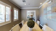 This business center offers a refined and functional workspace designed to support both collaborative meetings and individual productivity. The main conference room serves as the centerpiece, featuring a solid wood rectangular table surrounded by upholstered cream chairs, providing comfortable seating for multiple attendees. Warm taupe walls accented by crisp white crown molding and baseboards create a professional yet inviting atmosphere. Patterned carpeting in deep, neutral tones grounds the space with a sophisticated touch. Natural light filters in through plantation-shuttered windows, offering a bright and pleasant environment while maintaining privacy. Recessed lighting supplements the natural light, ensuring consistent illumination throughout the day. A decorative potted topiary centerpiece adds a subtle organic element to the conference table. French glass doors connect the conference room to a private inner office, which is visible in the background and equipped with a desk, lamp, and wall art — ideal for focused individual work or private consultations. The overall design balances practicality with aesthetic care, making this business center well-suited for professional meetings, collaborative work sessions, and quiet productivity.