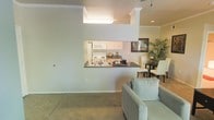 This single-floor residence offers a thoughtfully designed open-concept layout that maximizes both functionality and flow. The living area is carpeted and comfortably proportioned, providing a welcoming space for relaxation. A defining architectural feature is the pass-through kitchen window, which connects the kitchen to the main living and dining areas, creating an open and interactive feel throughout the home. The kitchen is equipped with white cabinetry, white appliances including a built-in microwave and range, and a dark countertop that extends to form a convenient bar-style ledge at the pass-through. Crown molding runs throughout the space, adding a refined touch to the interior. Adjacent to the kitchen, the dining area accommodates a table and chairs, complemented by lush indoor greenery that adds a natural, vibrant element to the space. The neutral wall tones and consistent flooring create a cohesive and calm aesthetic throughout. Recessed lighting and a ceiling-mounted light fixture provide ample illumination, contributing to the bright and airy ambiance of the home.