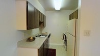 This single-floor space is designed with practicality and functionality in mind. The kitchen is laid out in a classic galley configuration, maximizing efficiency within a compact footprint. Dark wood cabinetry lines both sides of the kitchen, offering generous storage space through a combination of upper and lower cabinets. The light-toned countertops provide a clean contrast against the darker cabinetry and offer ample workspace for meal preparation. A built-in sink is seamlessly integrated into the counter, accompanied by a dishwasher nearby for added convenience. On the opposite wall, white appliances — including a freestanding range and a full-size refrigerator — complete the kitchen's essential offerings. The light tile flooring throughout the kitchen is easy to maintain and contributes to the room's bright, clean aesthetic. Overhead, a flush-mount fluorescent light fixture provides ample illumination across the workspace. Light-colored walls keep the interior feeling open and airy, while white baseboards add a crisp finishing touch. Overall, the space presents a straightforward, well-organized layout suited for comfortable, everyday living.
