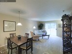 This single-floor residence features an open-concept layout that seamlessly connects the dining and living areas, creating a cohesive and functional living environment. The space is finished with wall-to-wall plush carpeting in a neutral tone, contributing to a warm and comfortable atmosphere throughout.

The dining area is anchored by a dark wood rectangular table paired with matching high-back chairs, grounded beneath a classic pendant light fixture that adds a focused, ambient glow. A framed artwork piece on the adjacent wall adds a subtle decorative touch.

The living area flows directly from the dining space and is furnished with a light-toned sofa and a coordinating armchair, arranged around a dark wood round coffee table. A floor lamp provides additional soft lighting, complementing the generous natural light that enters through a large sliding glass door fitted with vertical blinds. The glass door opens the room visually, offering an outdoor view and a bright, airy feel. A decorative potted plant in the corner adds a natural element, enhancing the overall calm and welcoming ambiance of the space.