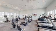 Located at 3465 Sandler Blvd in Little River, South Carolina, this comprehensive fitness center offers a generously sized, single-floor facility designed to support a full range of workout routines. The space is organized into several distinct zones, each tailored to a specific type of training.

The main cardio area is lined with TRUE-brand treadmills, ellipticals, and recumbent bikes, all arranged beneath ceiling fans and recessed lighting. Soft, neutral walls and multiple windows allow natural light to fill the room, creating a bright and energizing atmosphere. A wall-mounted television provides entertainment during workouts.

A dedicated strength and functional training zone features a fully stocked TRX-style training rig labeled "Training Zone," complete with suspension straps, resistance bands, barbells, weight plates, and medicine balls. Adjacent to this, a free weights area includes a dumbbell rack and adjustable benches alongside cable and cable-row machines.

A separate studio room offers a more intimate setting with hardwood-style flooring, a ballet barre, mirrored wall panels, a spin bike, and a rack of stability and medicine balls — ideal for stretching, yoga, or low-impact training.

The cycling and rowing area showcases floor-to-ceiling windows, soaring ceilings, and smart-screen-equipped spin bikes and rowing machines bathed in natural light. The facility also includes a clean, ADA-accessible restroom with classic hexagonal tile flooring and wainscoting.