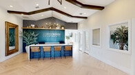The Copperwood Apartments clubhouse is a thoughtfully designed single-floor community space that seamlessly blends comfort, functionality, and modern style. The interior is anchored by striking vaulted ceilings adorned with exposed dark wood beams, creating an airy, welcoming atmosphere throughout. Light-toned herringbone hardwood flooring runs consistently across the entire space, lending an elegant, cohesive aesthetic.

At the heart of the clubhouse is a generously sized living area featuring a bold, floor-to-ceiling dark tile fireplace surround with a warm wood mantle and mounted flat-screen television. Deep teal wainscoting accents the lower walls, adding a rich, contemporary contrast to the crisp white upper walls and ceiling. Comfortable sectional seating, layered rugs, and curated décor create an inviting lounge environment.

The reception area makes a strong first impression with its navy blue bar-style front desk, woven rattan barstools, and a dramatic teal accent wall bearing the Copperwood Apartments branding beneath a stylish chandelier. Plantation shutters on the windows allow for controlled natural light throughout.

A dedicated co-working zone, highlighted by a sage green accent wall with display shelving, offers dual workstations with ample desk space. Adjacent to the lounge area, a game room and a kitchenette provide additional amenity spaces. Recessed lighting throughout ensures the entire clubhouse is bright and welcoming at all hours.