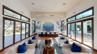 Located at 15400 W Goodyear Blvd N in Goodyear, Arizona, this single-floor community clubhouse offers a refined and welcoming gathering space designed with both comfort and style in mind. The interior is anchored by soaring vaulted ceilings and an abundance of natural light, which pours in through expansive floor-to-ceiling windows and multiple sets of glass doors that frame views of the surrounding outdoor amenities, including a playground and landscaped grounds. Warm terracotta tile flooring runs throughout, complementing the crisp white walls and dark-framed window trim for a clean, modern-Southwest aesthetic. The main lounge area features generously sized white upholstered seating accented with bold cobalt blue throw pillows, centered around a sculptural wood coffee table and layered area rugs. At the far end of the room, a dramatic arched alcove houses a mosaic tile backsplash in a sweeping gradient of aqua and deep blue, serving as the artistic centerpiece behind a sleek bar and countertop with white barstools. Elegant gold branching chandeliers illuminate both the lounge and bar areas, adding a touch of sophistication. Multiple glass door entries connect the interior to adjacent outdoor spaces, reinforcing the seamless indoor-outdoor flow that defines this sun-drenched Arizona retreat.