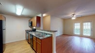 Located at 7461 Kitty Hawk Rd in Converse, TX, this single-floor residence offers a practical and comfortable layout designed for everyday living. The galley-style kitchen is well-appointed with warm wood cabinetry, dark granite-style countertops, black appliances including a range, microwave, and refrigerator, and a double-basin sink. A half-wall peninsula separates the kitchen from the open living and dining area, creating a connected yet defined space. The living area features durable wood-style flooring that extends throughout the main areas, neutral walls, and a ceiling fan with light fixture. A glass door provides natural light and outdoor access from the living space. The bedroom is generously sized with plush carpeting, multiple windows that invite natural light, and its own ceiling fan. The bathroom is clean and functional, featuring a combined tub and shower with tiled surround accents, a wood-toned vanity with granite-style countertop, and matching wood-style flooring. Throughout the unit, the consistent use of warm tones in the cabinetry, countertops, and flooring creates a cohesive and inviting atmosphere.