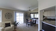 This single-floor residence offers a well-designed layout that balances comfort and functionality. The dining area serves as a central gathering space, featuring plush carpeting, crown molding, and warm neutral-toned walls that create an inviting atmosphere. A ceiling fan with a classic design keeps the space comfortable year-round while adding a subtle decorative touch.

Natural light pours in through large sliding glass doors that open to an outdoor patio area, offering pleasant exterior views and a seamless indoor-outdoor connection. The dining area comfortably accommodates a full dining set, making it ideal for everyday meals and entertaining alike.

A large framed mirror adorns one wall, adding depth and brightness to the room. Decorative indoor plants are thoughtfully placed throughout, enhancing the space with a touch of greenery. An open pass-through connects the dining area to the adjacent kitchen, which features dark cabinetry with stone countertops, promoting an easy flow between the two spaces.

The open-concept design ensures that the dining and living areas feel expansive and connected, while the clean white trim and crown molding throughout lend a polished, cohesive finish to the overall interior aesthetic.