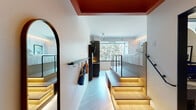 This thoughtfully designed studio maximizes every inch through clever split-level architecture. The entry zone features a bold rust-red ceiling accent, an arched mirror, and a wall-mounted coat rack, establishing a distinct arrival experience. The lower level houses a sleek kitchenette finished in dark cabinetry, complete with a black sink, induction cooktop, open shelving with warm underlighting, and a wall-mounted TV. Light wood flooring runs throughout, complemented by LED strip lighting that illuminates the base of the raised platform.

The bedroom occupies an elevated platform accessed via wide, LED-lit wooden steps with integrated storage beneath. Black metal railings define the platform's edge, adding an industrial contrast to the warm wood tones. A large picture window frames lush treetop views, flooding the sleeping area with natural light and creating a calm, immersive backdrop. Built-in cabinetry lines the walls for ample storage.

The bathroom is a design highlight, featuring bold cobalt blue wall tiles, black fixtures, a wall-hung toilet, and a glass-enclosed shower. A patterned tile floor and matte black framing complete the striking, contemporary aesthetic throughout the space.