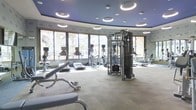 This fitness center showcases a striking curved architectural layout, creating a dynamic and open atmosphere that sets it apart from a standard gym environment. The ceiling is finished in a deep blue tone and features a series of circular recessed lighting fixtures that cast a clean, even illumination throughout the space, complementing the natural light that pours in through a continuous band of large floor-to-ceiling windows along the perimeter. These windows offer views of palm trees and the surrounding exterior, enhancing the bright and energizing ambiance of the facility. The flooring consists of neutral, durable carpet tiles well-suited for a high-traffic fitness environment. The equipment selection is comprehensive and modern, including multiple treadmills, elliptical cross-trainers, and a multi-station cable and strength training machine, as well as free weight benches — catering to a wide range of fitness routines. Mirrors are strategically positioned to maximize the sense of space and allow users to monitor their form during workouts. The overall design balances functionality with a polished, upscale aesthetic, making this fitness center a premium amenity suited for residents or guests seeking a well-appointed space for their exercise needs.