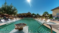 This expansive outdoor amenity complex is designed with a Mediterranean-inspired aesthetic, anchored by a large, crystal-clear swimming pool as its centerpiece. The pool features a decorative fountain feature and in-water lounge shelves lined with white sun chairs, ideal for relaxing in the sun. A separate spa provides an additional option for leisure and relaxation. Surrounding the pool, a generously sized deck is outfitted with multiple lounge chairs and shaded canopy umbrellas, offering both sun and shade options for guests. A substantial wood-beamed covered pavilion sits adjacent to the pool area, furnished with comfortable outdoor seating and dining sets, making it well-suited for gatherings and outdoor entertaining. Lush, mature landscaping throughout — including towering palm trees, shade trees, flowering plants, and manicured shrubs — creates a tropical, resort-like atmosphere. Neatly maintained green lawns and paved walkways connect the various areas of the complex. The clubhouse building, visible in the background and featuring a clay tile roof and stucco exterior, completes the cohesive Mediterranean character of the property. The entire outdoor space is bathed in abundant natural sunlight, enhancing its inviting, open-air ambiance.