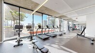 This contemporary fitness center is designed with both functionality and aesthetics in mind, delivering a premium workout environment. The space is defined by dramatic floor-to-ceiling glass walls that flood the interior with natural light and offer sweeping views of the surrounding cityscape, creating an energizing atmosphere for exercise.

The layout is thoughtfully organized across a single level, with dedicated zones for various types of training. The cardio section features a row of True-brand treadmills and stationary bikes with integrated screens, all positioned to take full advantage of the outdoor views. A rowing machine and flat benches complement this area for cross-training versatility.

The strength training section is equally well-appointed, housing a full cable and selectorized machine circuit, a power rack loaded with a colorful Olympic plate set, a comprehensive dumbbell rack, and a variety of resistance machines including a leg press and specialty functional trainer. A glass-partitioned room provides a more focused environment for targeted exercises.

Architectural details include exposed concrete ceiling elements contrasted against crisp white walls, linear pendant lighting, and durable speckled rubber flooring throughout. Direct access to an outdoor pool area is available via large glass doors, making this fitness center a well-rounded amenity space.